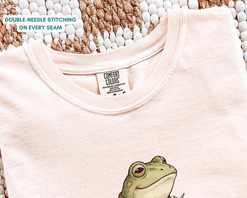 May include: A light peach-colored t-shirt with a crew neck and a frog graphic. The shirt has the text "DOUBLE-NEEDLE STITCHING ON EVERY SEAM" and a Comfort Colors tag. The frog is green with a brown collar.