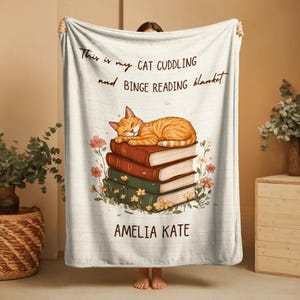 May include: A cream blanket with a design of a ginger cat sleeping on books. The blanket has the text "This is my cat cuddling and binge reading blanket" and the name "Amelia Kate".