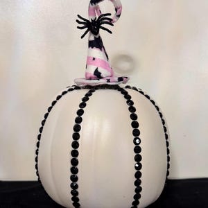 May include: A white pumpkin decorated for Halloween. The pumpkin is adorned with black rhinestones and topped with a witch's hat in pink, black, and white, with a black spider on top. A festive and decorative item.