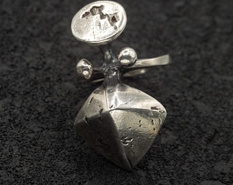 May include: A silver ring with an abstract design. The ring features a geometric, faceted shape at the base, a cluster of small spheres, and a circular top with an etched pattern. The ring is set against a dark, textured background.