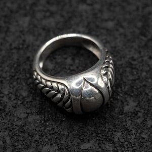 May include: A silver ring with a detailed design. The ring features a rounded, textured band with a central, raised element. The design includes rope-like detailing on the sides, creating a unique and eye-catching piece of jewelry.