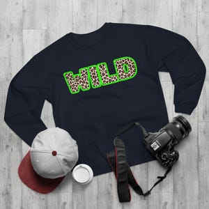 Puede incluir: Sudadera azul marino con la palabra "WILD" en letras grandes y audaces. Las letras están rellenas con un estampado de leopardo y delineadas en verde brillante. También se ven una gorra de béisbol y una cámara.