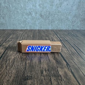 May include: A rectangular, tan-colored 3D printed object shaped like a candy bar wrapper. The front features a prominent blue and white label with the text SNICKERS in bold blue letters, outlined in white and red, set against a tan background.