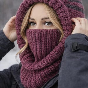 May include: A knitted hood and cowl set in a deep burgundy colour, with a ribbed texture on the cowl and a striped pattern on the hood. The hood covers the head and neck, with the cowl extending to cover the lower face.