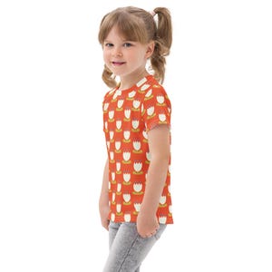 May include: A child's orange t-shirt featuring a repeating pattern of white and yellow floral designs. The short-sleeved top is made of a soft fabric and is worn by a young child. The shirt is paired with grey trousers.