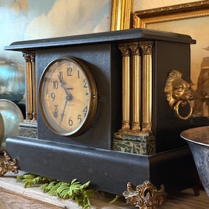 May include: A black mantel clock featuring a circular face with black Arabic numerals and hands, flanked by gold-toned fluted columns on green marble-like bases. The clock includes gold lion head side handles and ornate gold-toned metal feet.