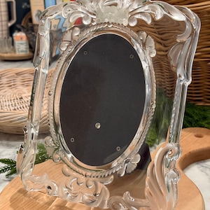 May include: A clear glass picture frame featuring an oval center opening and an ornate, molded border with decorative scrollwork and floral motifs. The frame includes a dark, solid oval backing insert held in place by four small metal pins.