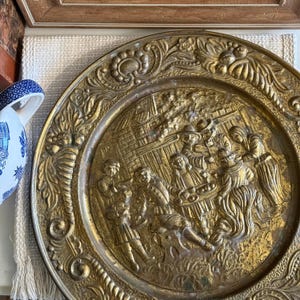 Könnte beinhalten: Ein runder dekorativer Messingteller mit einer detaillierten Prägung einer Gruppe von Menschen, die um einen Tisch versammelt sind. Der breite Rand ist mit komplexen Blumen- und Schnörkelmustern verziert. Das Metall hat ein warmes, gealtertes Goldfinish.