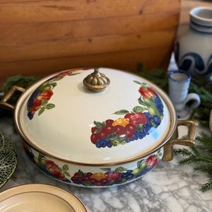 May include: A round, white enameled metal casserole dish with a matching lid, featuring a colorful fruit pattern of red cherries, blue berries, and green leaves. It has two brass-toned metal side handles and a brass-toned knob on the lid.