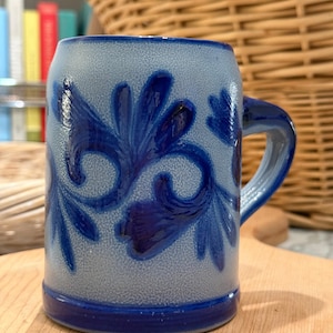 May include: A cylindrical ceramic mug with a light blue, textured salt-glaze finish. The mug features a dark blue, hand-painted floral pattern with swirling leaves and petals. It has a thick, dark blue rim and a matching dark blue base band.