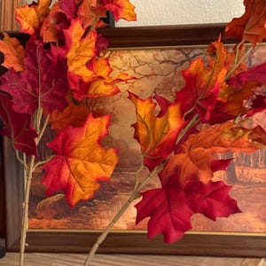 May include: Two artificial stems featuring vibrant autumn-coloured maple leaves in shades of deep red, bright orange, and golden yellow. The leaves have realistic veining and serrated edges, attached to thin, light-coloured, segmented plastic branches.