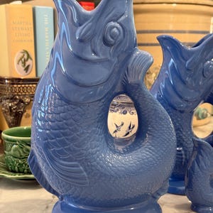 Op de afbeelding: Een glanzende blauwe keramische kan in de vorm van een vis met een open mond, gedetailleerde schubben en een gebogen staart die het handvat vormt. De kan staat op een platte ovale voet en heeft een gladde, gelijkmatige afwerking.