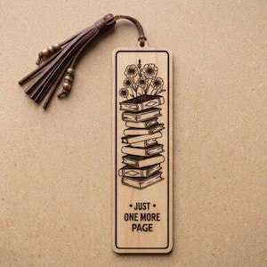 Puede incluir: Marcap&aacute;ginas de madera con un grabado de una pila de libros con flores. El marcap&aacute;ginas tiene el texto "JUST ONE MORE PAGE" y una borla de cuero marr&oacute;n. El marcap&aacute;ginas es rectangular con esquinas redondeadas.