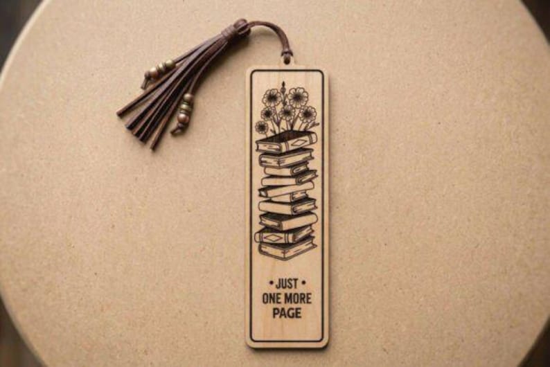 Puede incluir: Marcap&aacute;ginas de madera con un grabado de una pila de libros con flores. El marcap&aacute;ginas tiene el texto "JUST ONE MORE PAGE" y una borla de cuero marr&oacute;n. El marcap&aacute;ginas es rectangular con esquinas redondeadas.