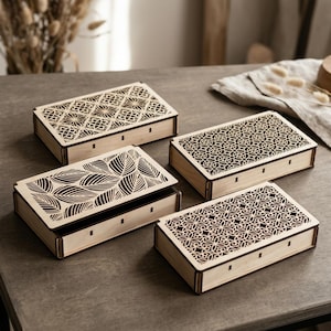 Può includere: Quattro scatole rettangolari in legno chiaro con coperchi scorrevoli. Ogni coperchio presenta un motivo unico tagliato al laser: uno con design geometrico a stella, uno con motivo a foglie e due con complessi reticoli floreali.