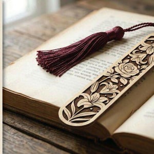 Puede incluir: Un marcap&aacute;ginas de madera cortado con l&aacute;ser con dise&ntilde;os florales, incluyendo rosas y lirios, y una borla burdeos. El marcap&aacute;ginas est&aacute; colocado en un libro abierto con p&aacute;ginas envejecidas. El texto "LASER CUT BOOK MARK" es visible.
