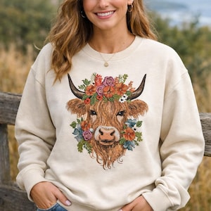 May include: A cream-coloured sweatshirt with a Highland cow design, decorated with a floral crown. The cow has brown fur, black horns, and is surrounded by colourful flowers. The sweatshirt is a casual, comfortable style.