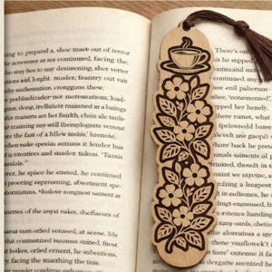Puede incluir: Marcap&aacute;ginas de madera cortado con l&aacute;ser con un dise&ntilde;o floral y una ilustraci&oacute;n de taza de caf&eacute;. El marcap&aacute;ginas tiene una borla marr&oacute;n y se muestra en un libro abierto. El texto "LASER CUT BOOK MARK" es visible.