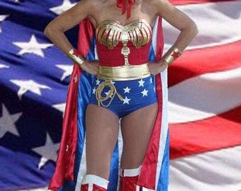 May include: A woman dressed as Wonder Woman stands in front of an American flag. She is wearing a red, blue, and gold costume with a star-spangled skirt and a red cape. She is also wearing a gold tiara with a red star on top and red boots with white accents.