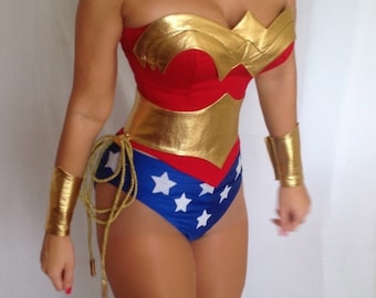 May include: A Wonder Woman costume featuring a red and gold bodice, blue shorts with white stars, and gold wrist cuffs. The costume has a strapless design and a gold belt. A gold rope is tied around the waist.