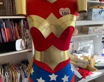 May include: A red and gold Wonder Woman costume with a blue skirt and white stars. The costume is on a dress form and includes a gold belt, chest piece, and arm cuffs. The dress form has a label that says "Singer 150".