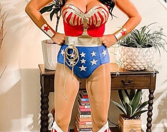 May include: A woman dressed as Wonder Woman, wearing a red, white, and blue costume with a gold belt and a red star on her headband. She is standing with her hands on her hips, wearing red boots with white accents.
