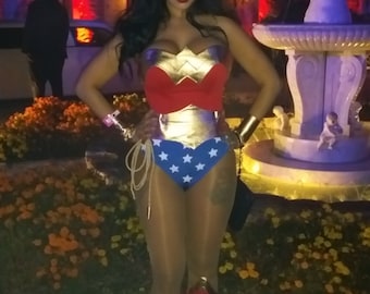 May include: A woman dressed as Wonder Woman, wearing a red, gold and blue costume with a star-spangled skirt and red boots. She is standing in front of a fountain with flowers in the background.