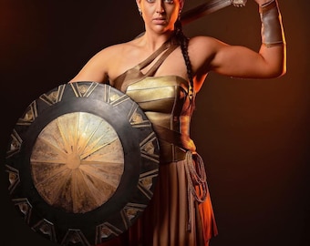 May include: A woman in a gold and tan costume, holding a sword and shield. The shield is black with gold accents and a sunburst design. The costume includes a corset, skirt, and arm cuffs. The background is dark.