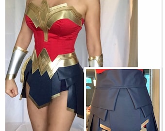 May include: A Wonder Woman costume featuring a red bodice with gold trim and a blue skirt with gold accents. The skirt is layered and has a pleated design.