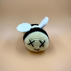 May include: A crocheted bee toy with a cream-colored body and black stripes. The bee has white wings and black stitched eyes in an "X" shape. The toy is handmade and sits on a solid, light orange background.