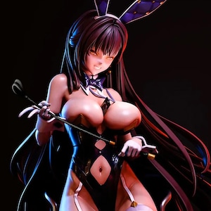 Op de afbeelding: Een beeldje van een vrouw in een zwart konijnenpak, compleet met konijnenoren en een zweep. De figuur poseert op een kruk, met lang donker haar en witte kousen. De belichting is dramatisch, met rode en blauwe tinten.