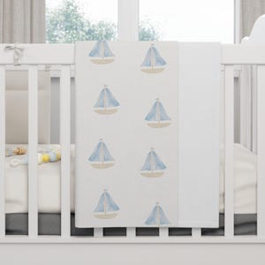 May include: A white baby blanket featuring a pattern of blue sailboats. The sailboats have light blue sails and beige hulls. The blanket is folded over a white cot.