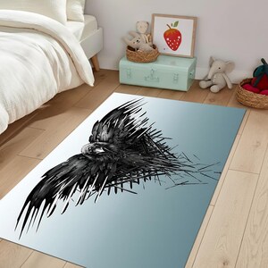 May include: A rectangular area rug featuring a black and white graphic of a raven in flight. The bird's wings are spread, and the design has a modern, artistic style. The rug has a gradient blue background.