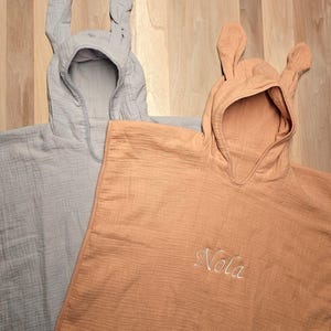 Könnte beinhalten: Zwei Baby-Badeponchos mit Kapuze und Hasenohren. Einer ist hellgrau, der andere pfirsichfarben und mit dem Namen "Nola" in Schreibschrift bestickt. Die Handtücher sind aus einem weichen, strukturierten Stoff.