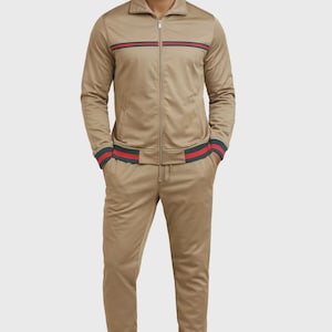 May include: Tan tracksuit with a zip-up jacket and matching pants. The jacket and pants feature red and green stripes. The cuffs and ankles have matching striped detailing. The outfit is worn with white sneakers.