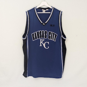 May include: A blue mesh jersey with the words "KANSAS CITY" in white arched lettering. The jersey has a V-neck with white and black trim, and the letters "KC" are visible. The jersey is hanging on a white hanger.