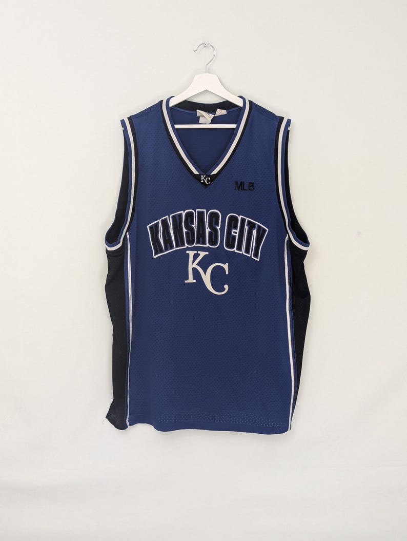 May include: A blue mesh jersey with the words "KANSAS CITY" in white arched lettering. The jersey has a V-neck with white and black trim, and the letters "KC" are visible. The jersey is hanging on a white hanger.