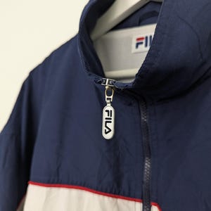 May include: A navy blue jacket with a white and red stripe across the chest. The jacket has a high collar and a zipper closure with the brand name "FILA" on the zipper pull. The jacket is hanging on a white hanger.