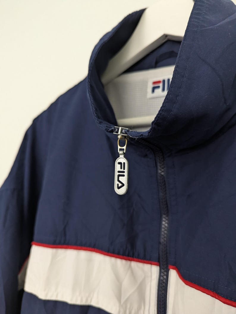 May include: A navy blue jacket with a white and red stripe across the chest. The jacket has a high collar and a zipper closure with the brand name "FILA" on the zipper pull. The jacket is hanging on a white hanger.