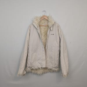 May include: Cream-colored Adidas jacket with a faux fur collar and lining. The jacket has a zip closure, side pockets, and a small Adidas logo on the chest. The cuffs and hem feature decorative buckles.