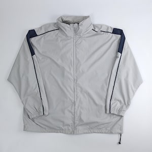 Pode incluir: Um blusão corta-vento cinza-claro com fecho frontal completo, gola alta e mangas compridas com painéis azul-marinho escuro ao longo dos ombros e braços. O casaco inclui punhos elásticos e uma bainha com cordão ajustável.