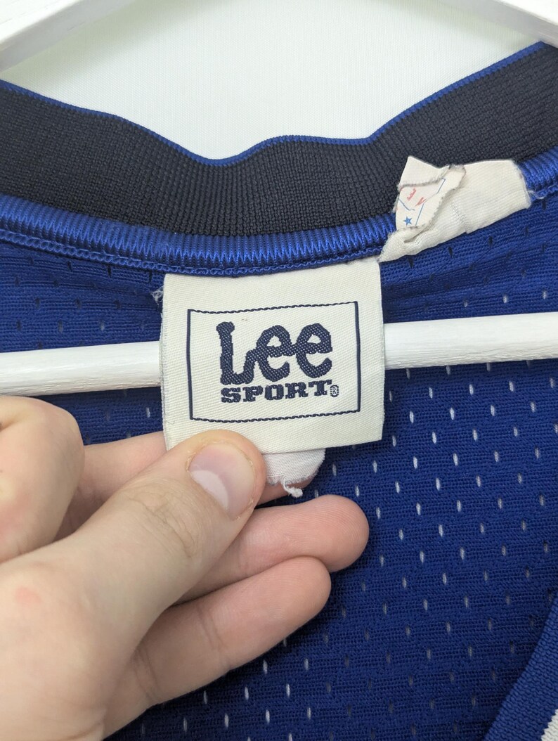 May include: Close-up of a blue mesh jersey with a navy blue collar. The jersey features a "Lee Sport" tag. The tag is white with black text. The jersey is hanging on a white hanger.