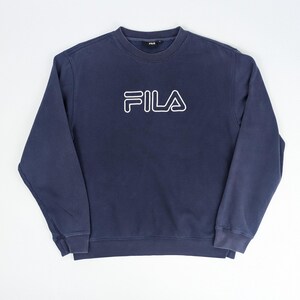 May include: A navy blue sweatshirt with the white outlined word "FILA" across the chest. The crew neck sweatshirt is a solid color and has long sleeves. The garment is made of a soft material.