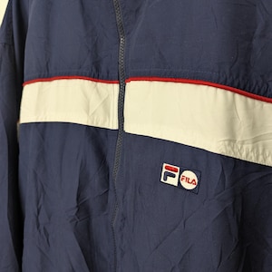 May include: Navy blue Fila jacket with a white stripe across the chest, accented with a thin red stripe. The jacket features a full zipper and the Fila logo on the chest and upper left shoulder. The jacket is made of a lightweight material.