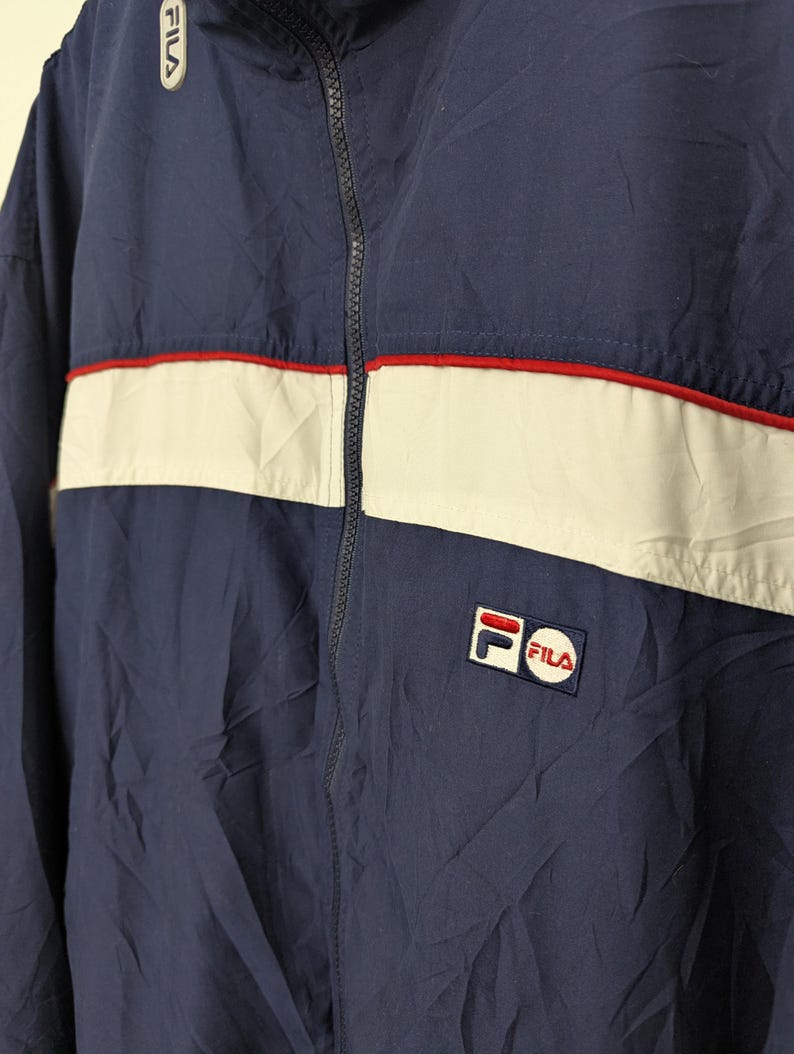 May include: Navy blue Fila jacket with a white stripe across the chest, accented with a thin red stripe. The jacket features a full zipper and the Fila logo on the chest and upper left shoulder. The jacket is made of a lightweight material.
