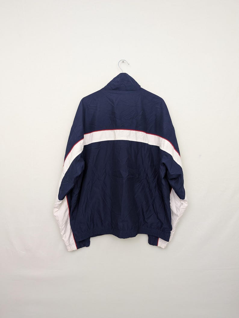 May include: Navy blue track jacket with white and red accents. The jacket features a full zipper, a high collar, and white stripes across the chest and sleeves. The cuffs and hem are finished with a red trim.