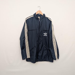 Op de afbeelding: Marineblauwe Adidas-trainingsjas met drie crèmekleurige strepen langs elke mouw en het Adidas-logo op de borst. De jas heeft een volledige ritssluiting en twee voorzakken. Hij hangt aan een houten hanger.