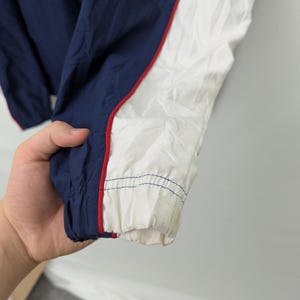 May include: Close-up of a navy blue and white jacket sleeve. The jacket has a red stripe along the white panel. The cuff is elasticized and shows signs of wear. The jacket is made of a lightweight, possibly nylon, material.
