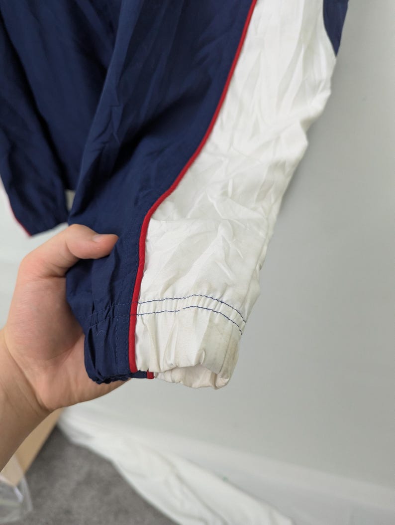 May include: Close-up of a navy blue and white jacket sleeve. The jacket has a red stripe along the white panel. The cuff is elasticized and shows signs of wear. The jacket is made of a lightweight, possibly nylon, material.