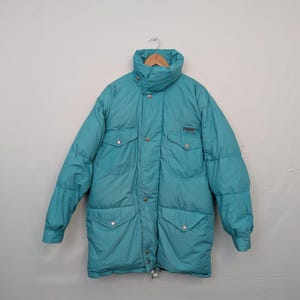 May include: A teal puffer jacket with a high collar and multiple pockets. The jacket features the Puma logo on the chest and is hanging on a wooden hanger. The jacket appears to be a vintage style.